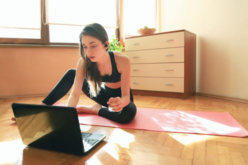 A person sits on their yoga mat and looks at their laptop to choose a workout video. Working out in ...