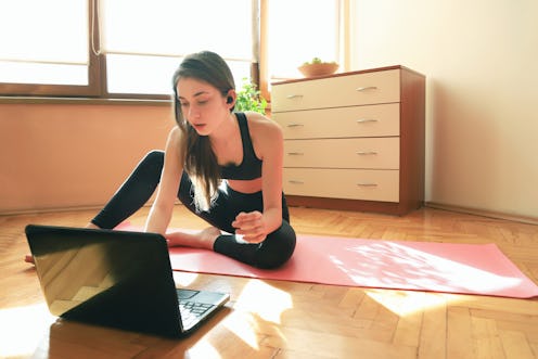 A person sits on their yoga mat and looks at their laptop to choose a workout video. Working out in ...
