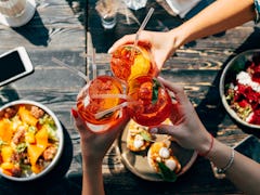 A group of friends toasts with glasses of Aperol Spritz, while eating a colorful meal in the summert...