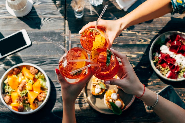 A group of friends toasts with glasses of Aperol Spritz, while eating a colorful meal in the summert...
