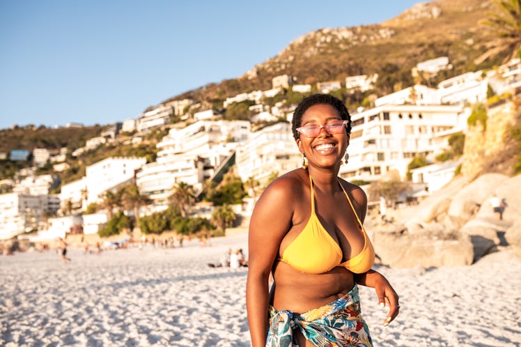 A young woman stands on the beach in a yellow string bikini and smiles.