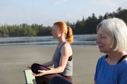 A mother and daughter sit cross-legged on their yoga mats on the beach.