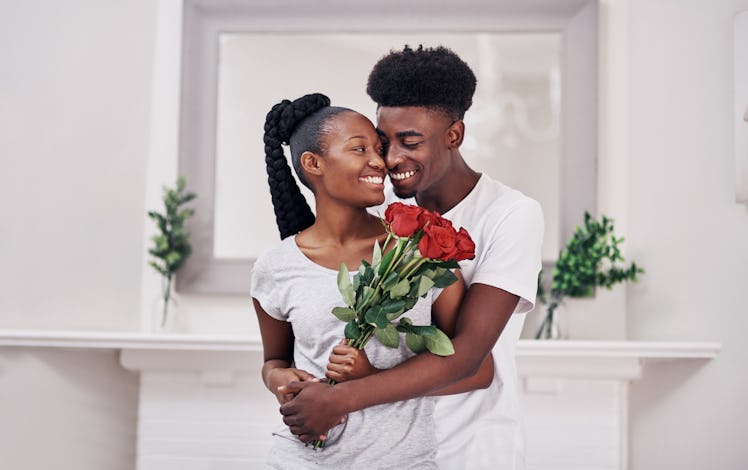 A young couple holds each other and a bouquet of roses while standing in their home.