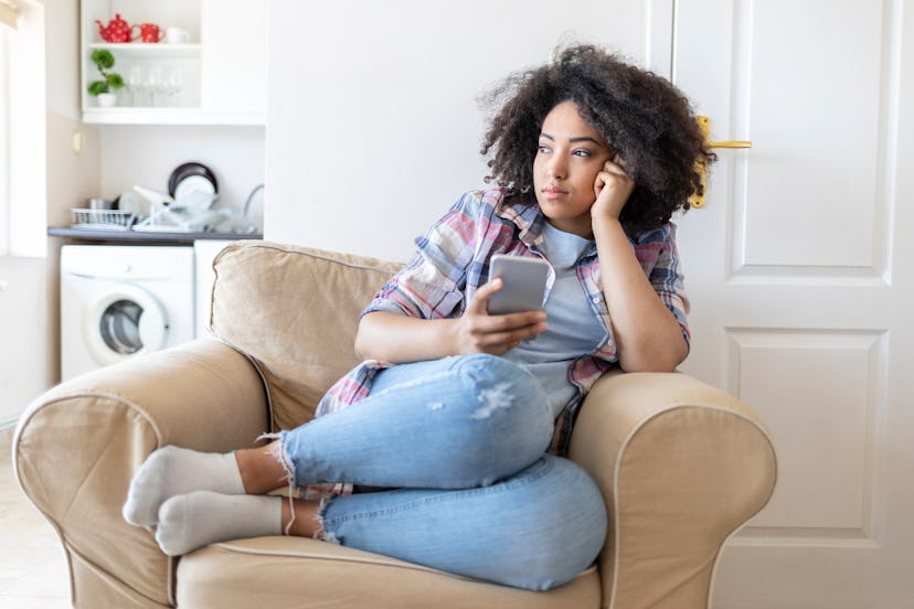 A curly-haired woman sitting in an armchair looking through the window and holding her phone while t...