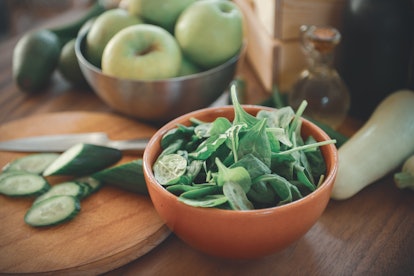 A bowl of spinach, next to a cutting board with sliced cucumber and a bowl of green apples blurred i...