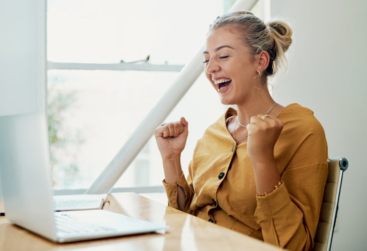 A young woman cheers while sitting at her kitchen table on a spring day and video chatting on her la...