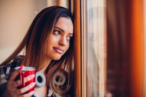 A brunette woman looking through her window whilst holding a red mug