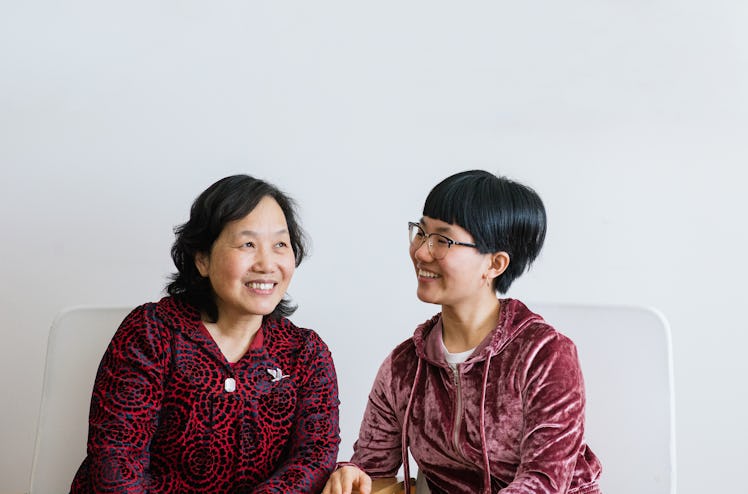 An older mom and daughter sit on a white couch and smile candidly.