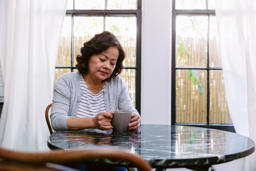 An older woman with Alzheimer sitting next to a window and having a cup of coffee