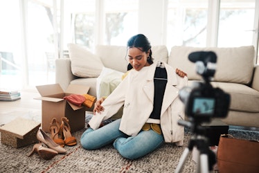 A young woman sits on her living room floor and unboxes new clothes while recording herself on a dig...