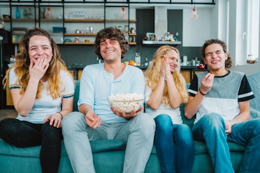 A family sits on a teal couch in their home and laughs while watching a movie and eating popcorn.