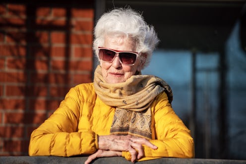 An old lady with Alzheimer sitting on a balcony wearing a yellow puffer jacket, sunglasses and a sca...