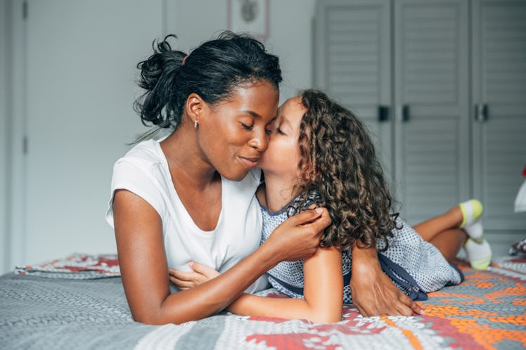 A young mom lays on her bed and hugs her daughter, who's kissing her on the cheek.