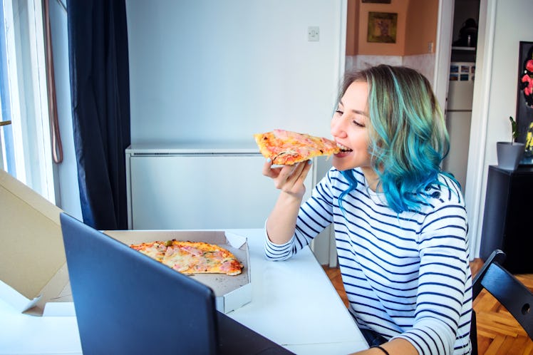 A young woman with blue-green hair plays an escape room on her laptop and eats a slice of pizza.