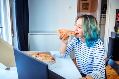 A young woman with blue-green hair plays an escape room on her laptop and eats a slice of pizza.