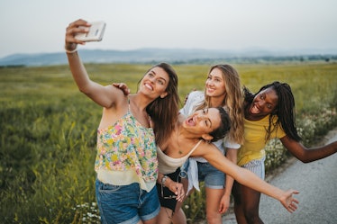 Four young women take a funny selfie while standing next to a grassy field.
