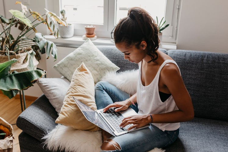 A young woman sits on a couch with her laptop and is surrounded by plants.