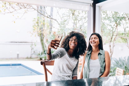 Two young women smile while sitting on an outdoor patio and taking a selfie.