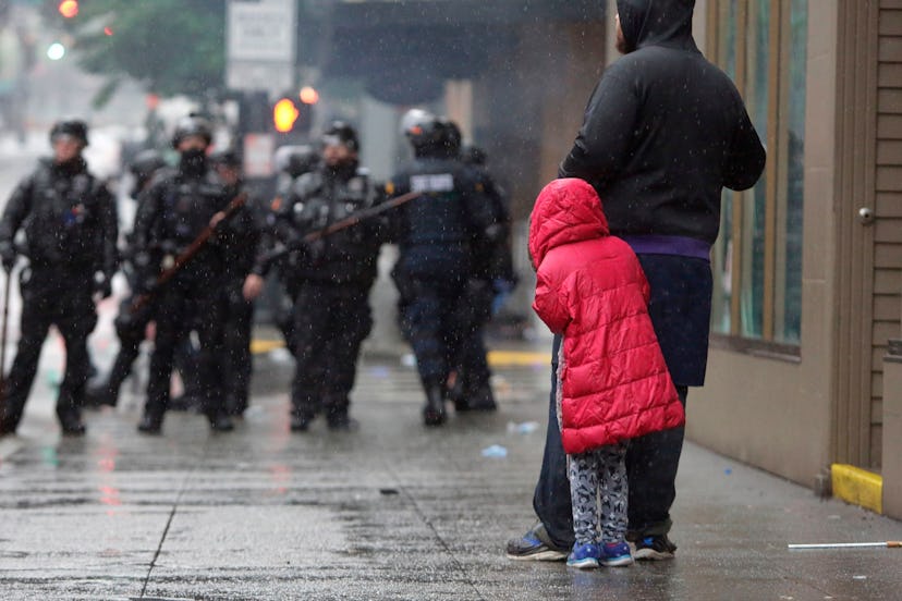 A childl hides behind an adult in front of a line of police