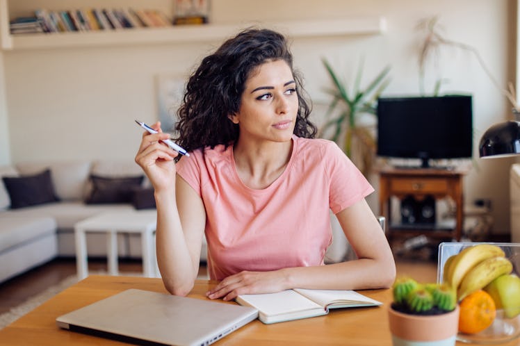 A young woman looks out the window while writing in a journal and sitting at her kitchen table.