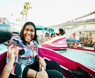A young Black woman laughs while riding in a hot pink cart at an amusement park in the summertime.