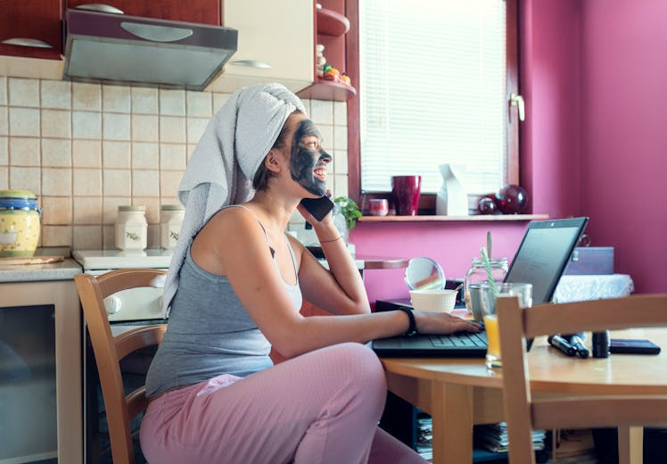 A young woman sits at her kitchen table and talks on the phone while doing a facial and wearing her ...