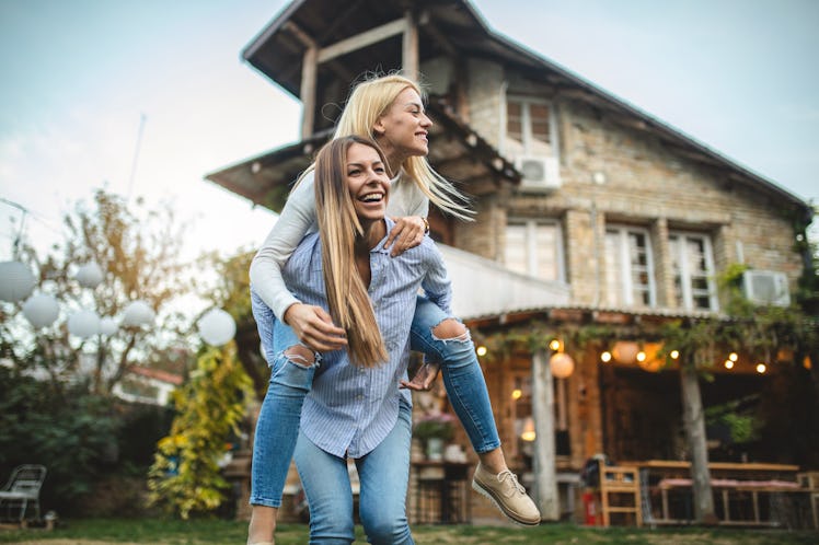 Two happy blonde friends piggyback and play around in the backyard in the summer.