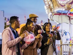 A Black family goes to a carnival at night and eats cotton candy and candy apples near a merry-'go-r...