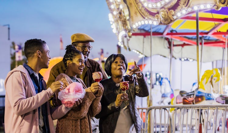 A Black family goes to a carnival at night and eats cotton candy and candy apples near a merry-'go-r...