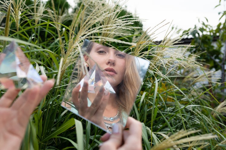 A young woman holds a glass prism and poses in a small mirror while sitting in a grassy field in the...