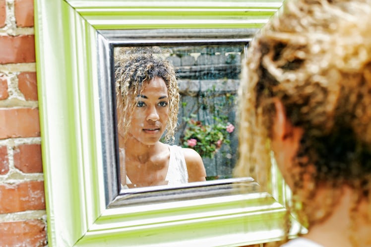 A young Black woman poses in a green-framed mirror that's on a brick wall outdoors.