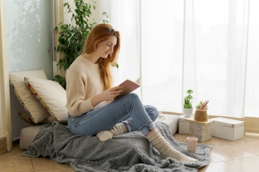 A woman sits in a comfy reading nook by her window with a cup, cozy socks, and a book. 