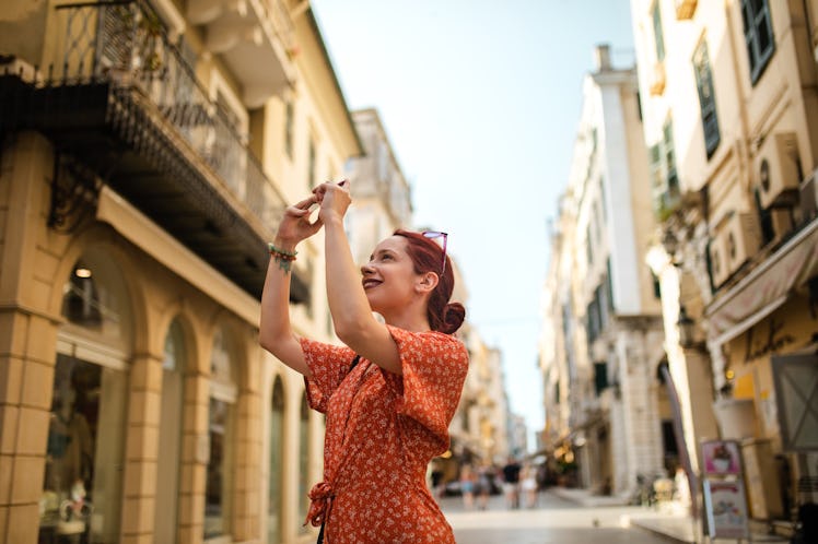 A young woman in a red dress takes a picture on her phone while vacationing in Greece.
