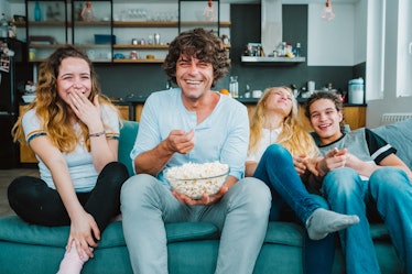 A group of siblings laughs with their dad while sitting on their couch and eating popcorn.