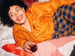 A young Black woman lays in bed with a glass of red wine on her birthday and smiles in a brightly-co...