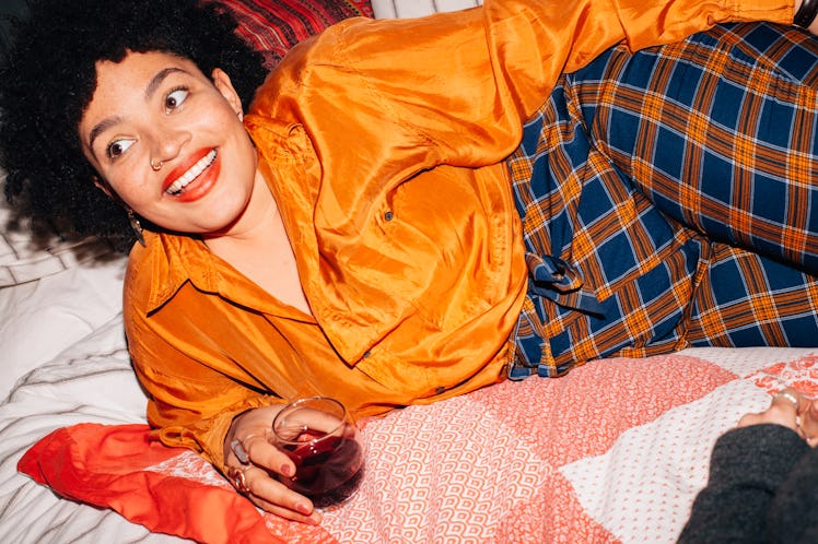 A young Black woman lays in bed with a glass of red wine on her birthday and smiles in a brightly-co...
