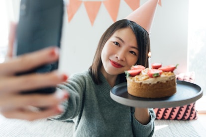 A young Asian woman poses with her birthday cake and takes a selfie on her phone.