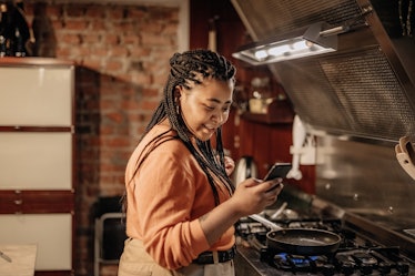 A woman looks at her phone, while standing next to her stove in the kitchen. 