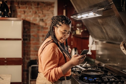 A woman looks at her phone, while standing next to her stove in the kitchen. 