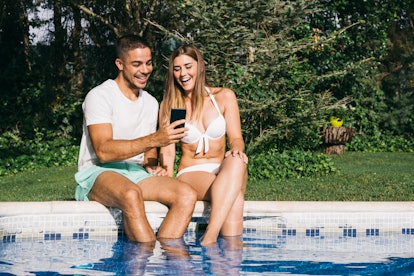 A laughing couple, sitting by the pool, looks at their phone. 