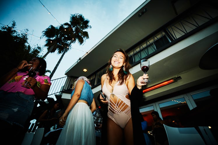 A young woman stands with her friends in a tropical destination with a glass of wine and a bathing s...