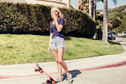 A young woman stands on the street in her neighborhood with a skateboard in the middle of summer.