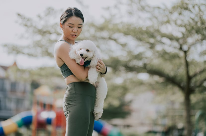 A person wearing dark green workout clothes holds their white puppy close to their chest in a park. ...