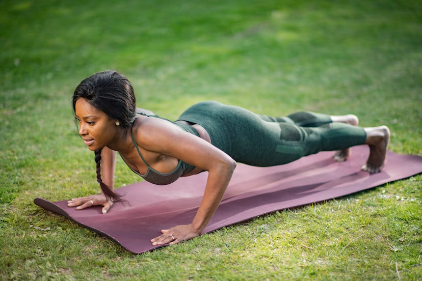 A woman does a plank on a purple yoga mat outside. If you're quarantining with your dog, doing worko...