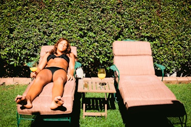 A young woman lays in a pink lounge chair in her backyard on a sunny day with a drink in her hand. 