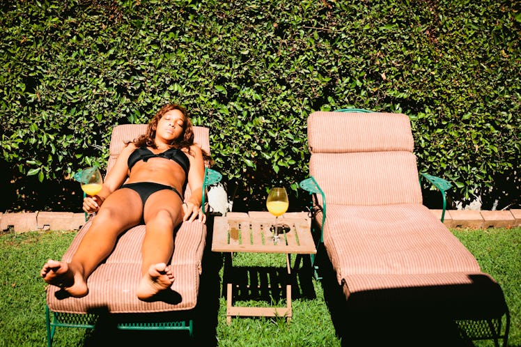 A young woman lays in a pink lounge chair in her backyard on a sunny day with a drink in her hand.