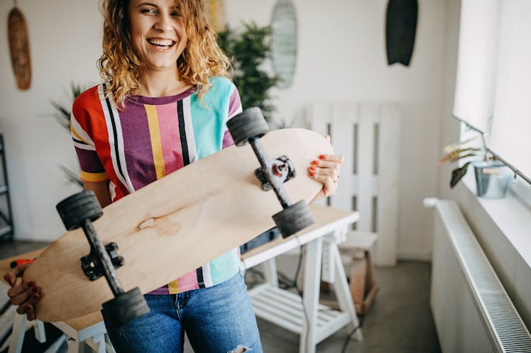 A young woman laughs while holding a skateboard in her home.