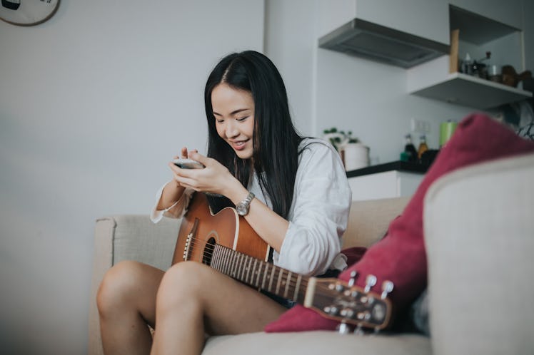 A young Asian woman holds a guitar on her lap while playing with her phone.