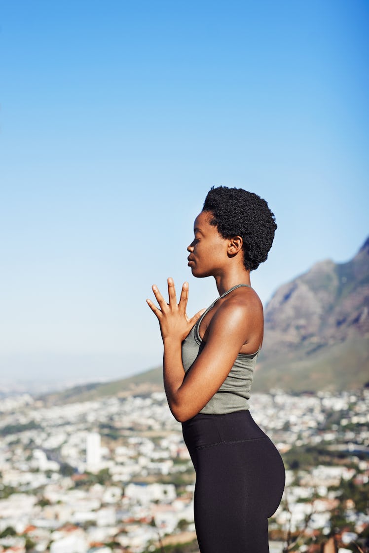 A young Black woman stands on a mountain overlooking a city and meditates on a sunny day.