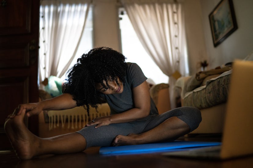 A person with natural hair wearing yoga pants sinks into a stretch on their yoga mat at home while t...